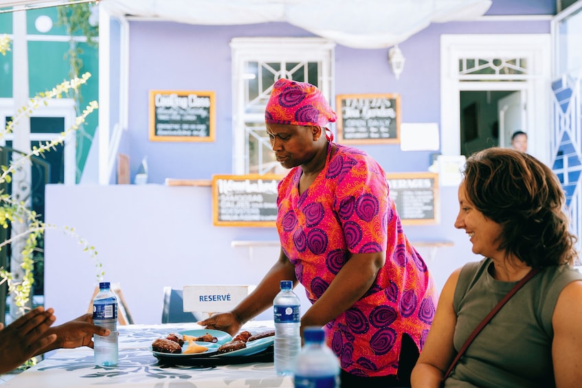 A person in a vibrant pink and purple patterned outfit serving food at a table with a 'RESERVED' sign. 4
