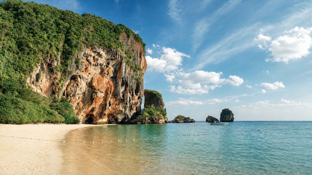 A beach with clear water, a large cliff, and a boat in the distance. 2