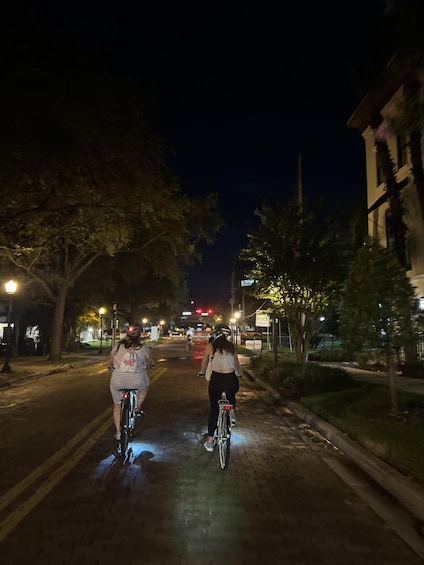 Two cyclists on a paved path at night, with streetlights and buildings in the background. 1