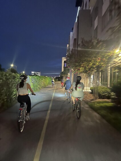 Cyclists on a paved path at night, flanked by buildings and greenery. 5
