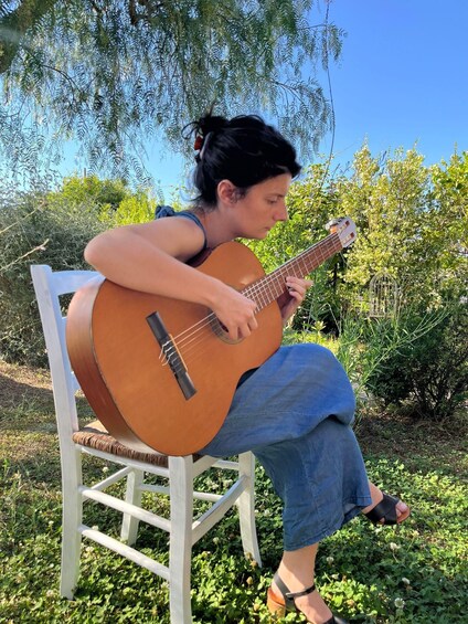 A person playing an acoustic guitar seated on a white chair outdoors. 2