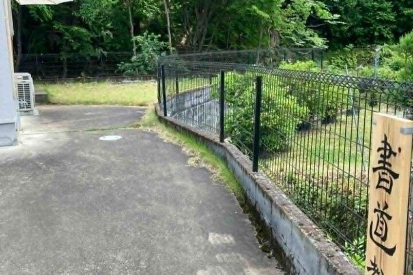A paved pathway leading to a fenced area with greenery and a wooden sign with Japanese characters. 4