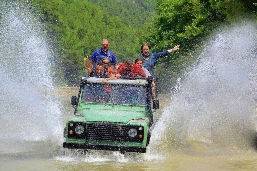 A green off-road vehicle driving through a shallow body of water with passengers. 5