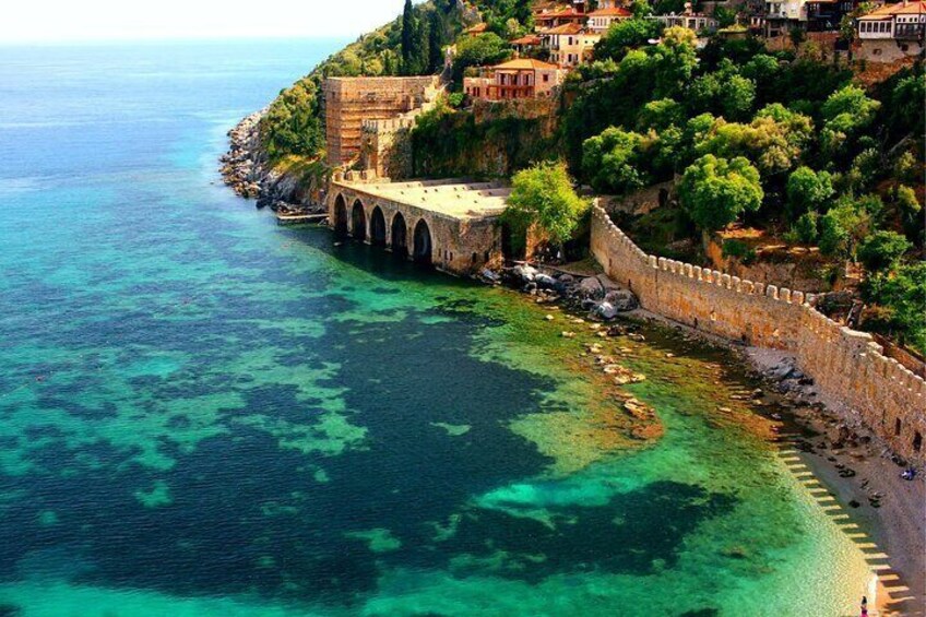 A coastal town with clear blue waters, a stone bridge, and buildings on a hillside. 1
