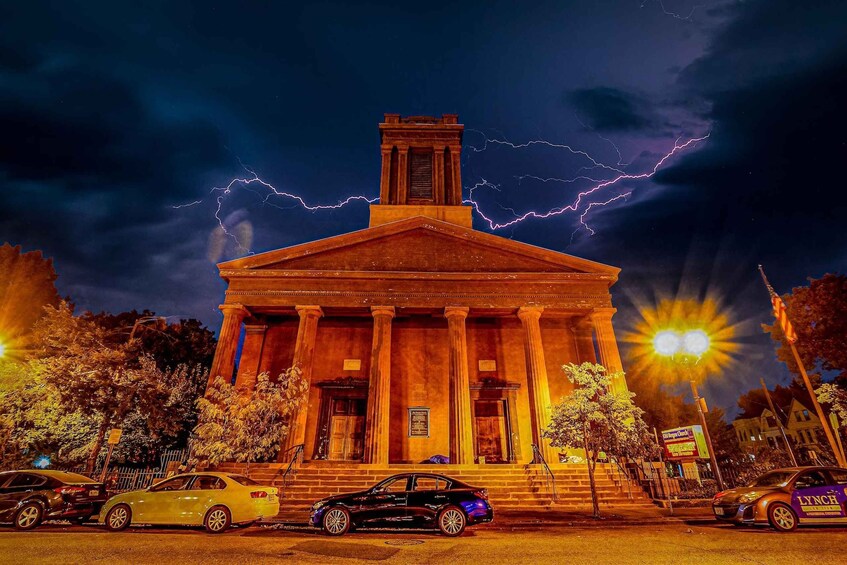 A neoclassical building with columns, parked cars, and lightning in the sky. 1