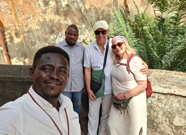 A group of people standing outdoors with a stone wall and greenery in the background. 1