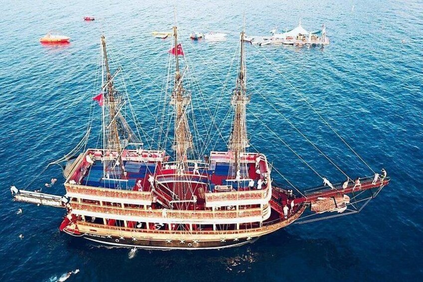 A large, multi-decked ship with multiple masts and a red and white color scheme, floating on a body of water with other smaller boats in the background. 4
