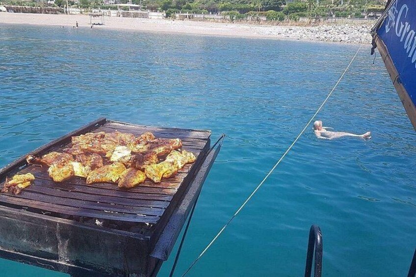 A grill with food on a boat, a person swimming in the water, and a beach in the background. 3
