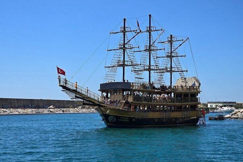 A tall ship with multiple decks and a Turkish flag, docked at a harbor with calm waters and rocky shore. 1