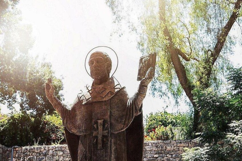 A statue of a person with a halo, holding a book and a tablet, standing in a garden with trees and a stone wall. 4