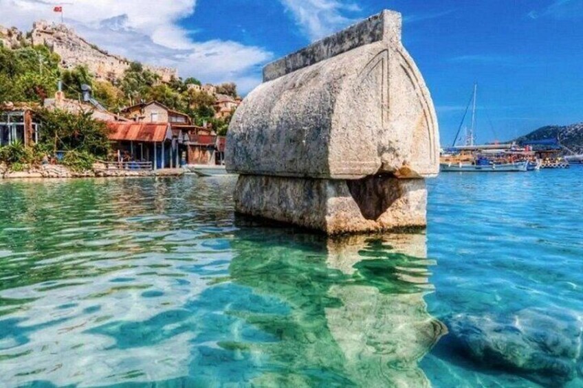 A clear blue sea with a stone structure in the water, a rocky hillside with buildings, and a clear sky. 1