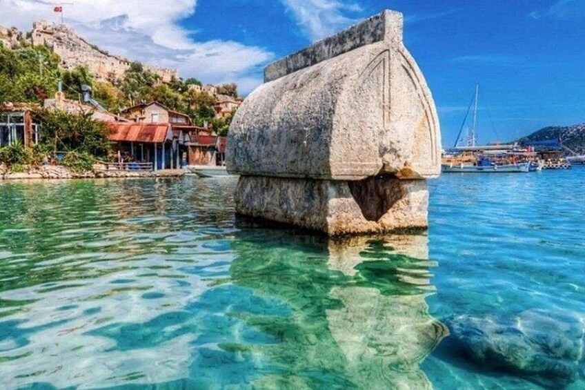 A clear blue sea with a stone structure in the water, a coastal town with buildings, and a hill with a flag. 1