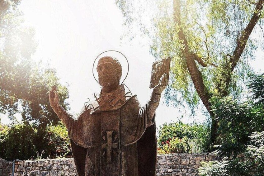 A statue of a robed figure with a halo, holding a book and a tablet, set against a stone wall and trees. 4