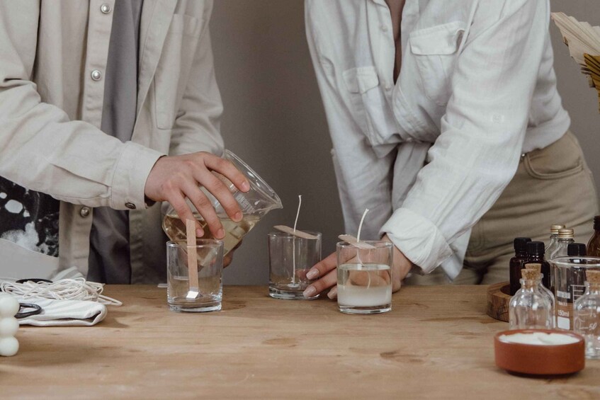 Two people preparing a mixture in small glass containers on a wooden table, surrounded by various bottles and a bowl. 1
