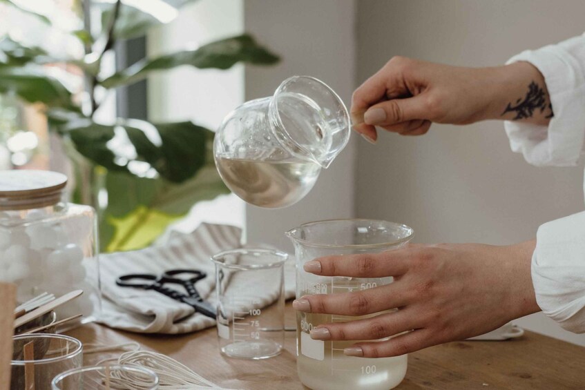 A person pouring a clear liquid into a beaker on a wooden table with measuring cups and a plant in the background. 4
