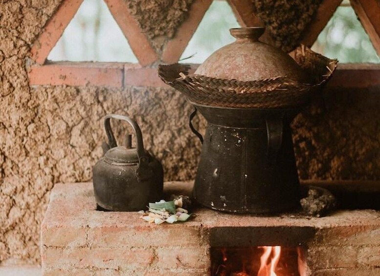 A rustic outdoor setting with a brick stove, a metal kettle, and a pot on a wooden table. 4