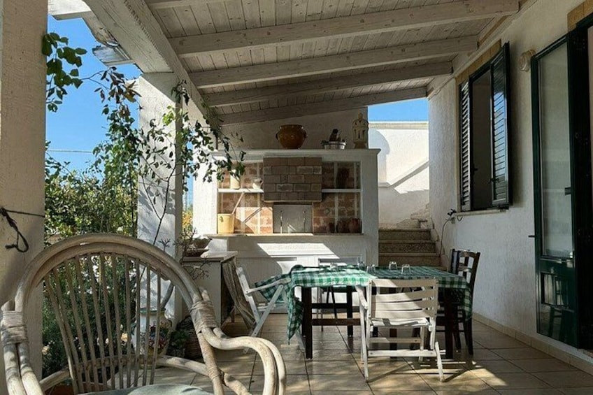 A covered outdoor dining area with a table and chairs, a bicycle, and a rustic kitchen in the background. 2