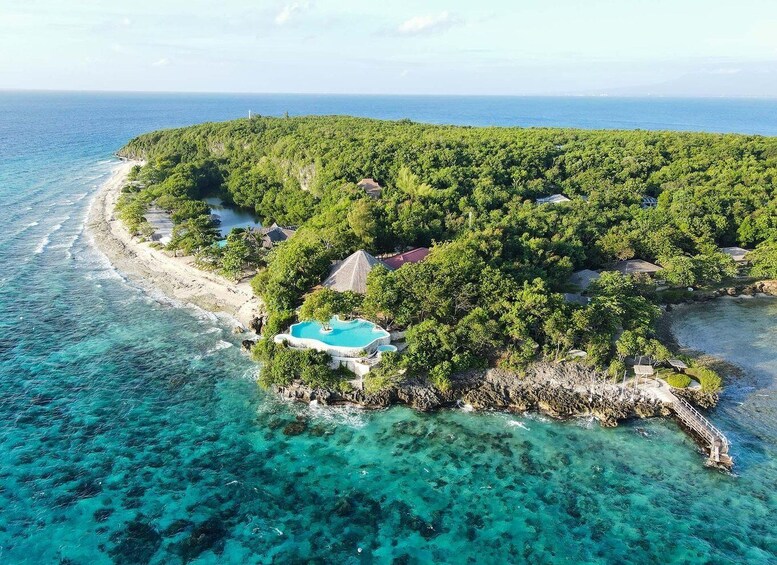 A tropical island with a clear blue sea, a small beach, and a swimming pool surrounded by lush greenery. 2
