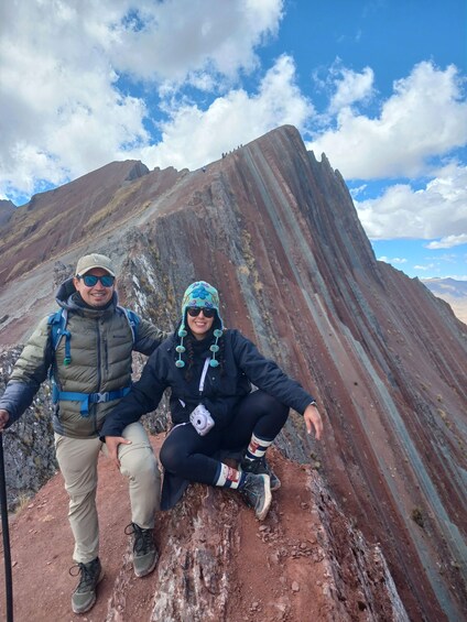 Two people hiking on a rocky mountain trail with a large, striped mountain in the background. 2