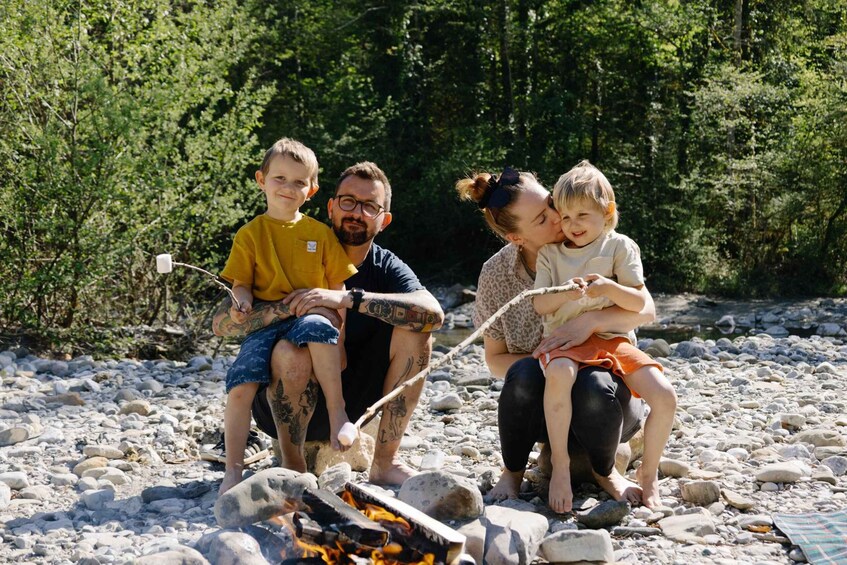 A family of four sitting around a campfire on a rocky beach with dense greenery in the background. 1