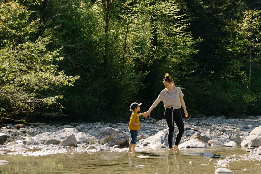 A woman and child holding hands by a river with rocks and trees in the background. 4