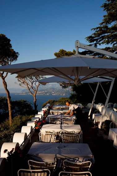 An outdoor restaurant with white tables and chairs, overlooking a scenic view of the sea and mountains. 2