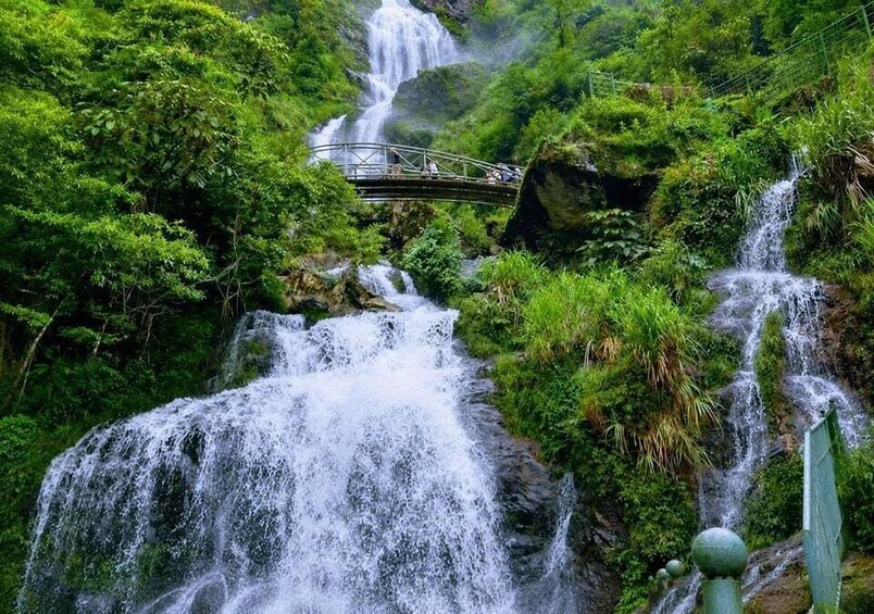 A multi-tiered waterfall with a bridge in a lush green forest. 5