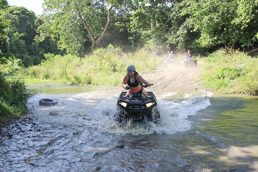 A person riding an ATV through a shallow river surrounded by greenery. 4