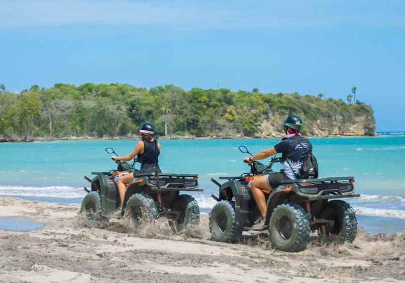 Two people riding ATVs on a beach with clear blue water and a forested island in the background. 1