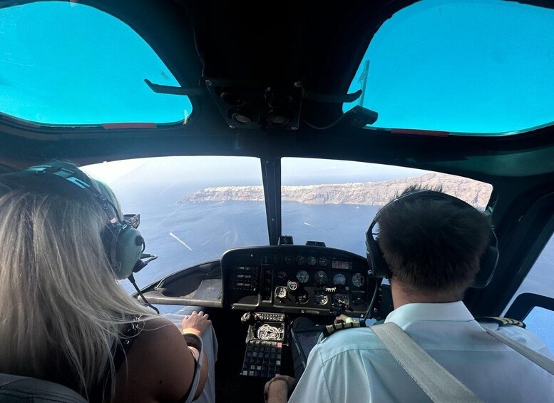 A helicopter cockpit with two people, one in the pilot's seat and one in the passenger seat, overlooking a coastline. 1
