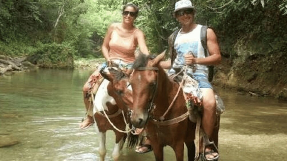 Two people riding a horse through a shallow river in a forested area. 4