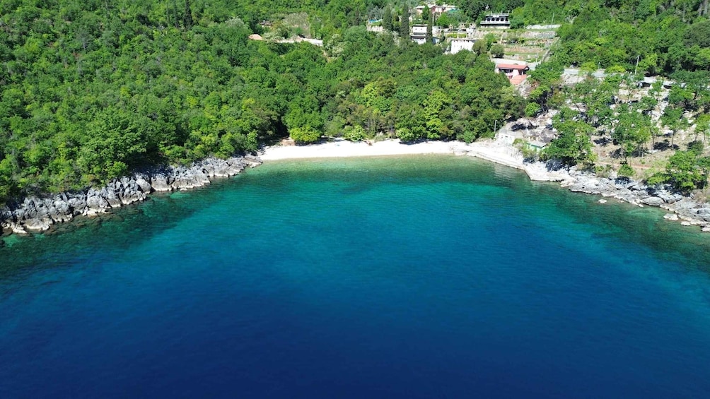 A coastal landscape with a clear turquoise sea, rocky shoreline, dense green forest, and scattered buildings. 1