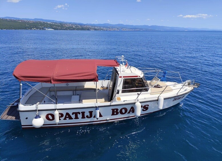 A white boat named 'OPATIJ BOA'S' with a red canopy, floating on a blue sea with a distant shoreline. 2