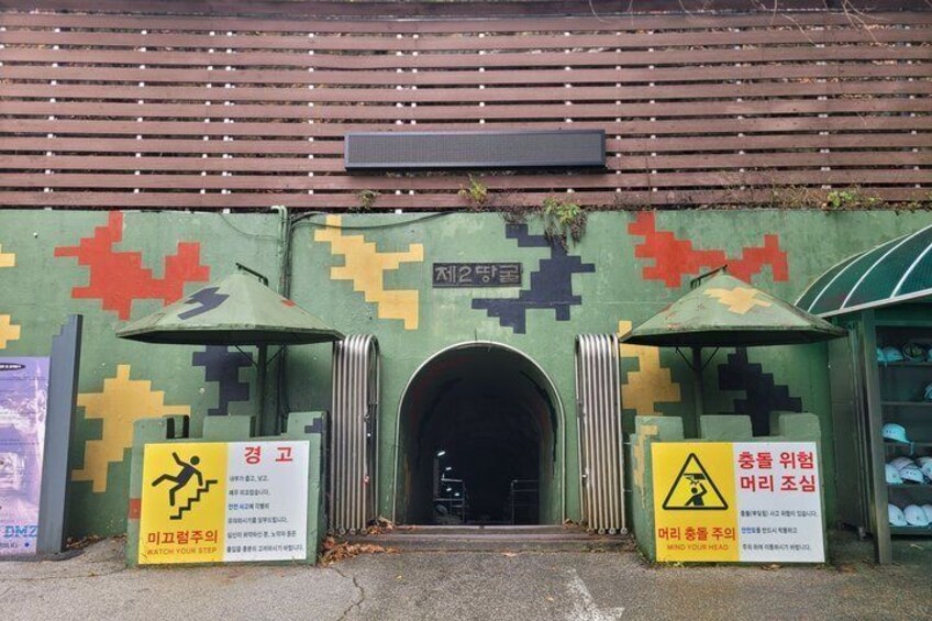 A building entrance with a camouflage-patterned wall, two warning signs, and a metal gate. 1