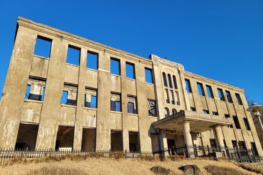 A large, multi-story building with a flat roof and multiple windows, surrounded by a fence and dry grass. 3