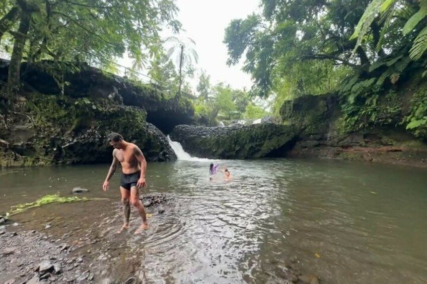 A person standing in a shallow river surrounded by lush greenery and a waterfall in the background. 3