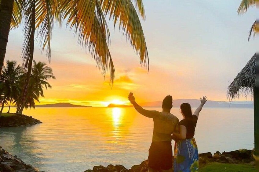 A sunset over a calm sea with palm trees and a couple embracing. 1