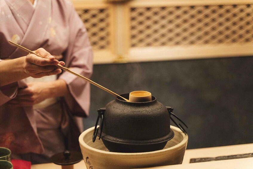 A person in a traditional robe holding a chopstick over a black pot on a wooden stand. 4