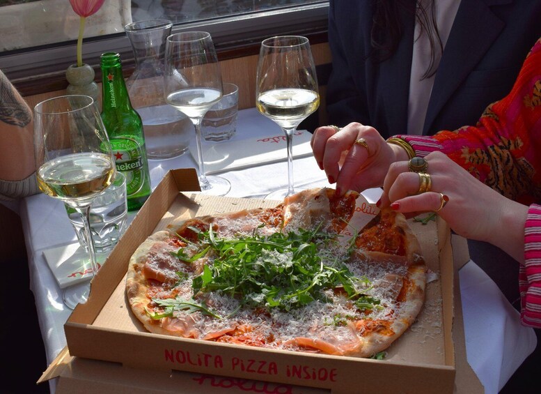 A pizza box labeled 'NOLITA PIZZA INSIDE' with a pizza topped with greens and cheese, a bottle of Heineken, and wine glasses on a table. 1