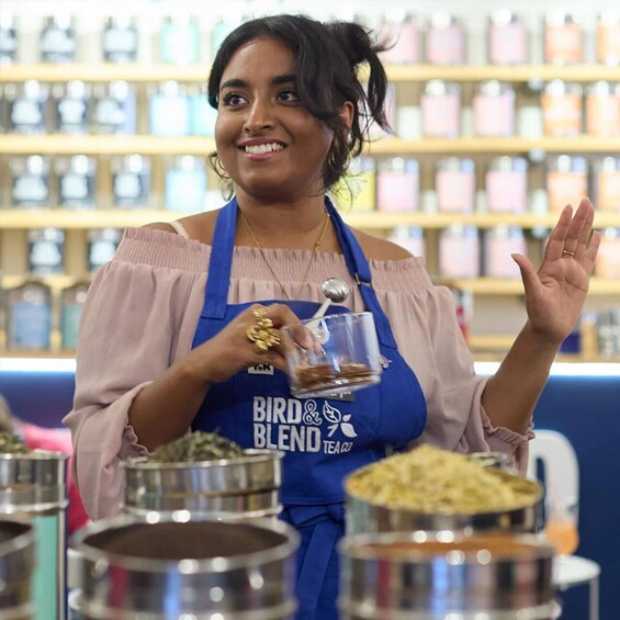 A person in a blue apron with 'BIRD & BLEND TEA' branding, holding a glass with a spoon, standing in front of multiple stainless steel containers with various contents. 3