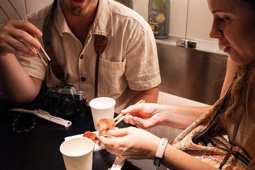 Two people eating pizza at a table with a camera and a bottle in the background. 1