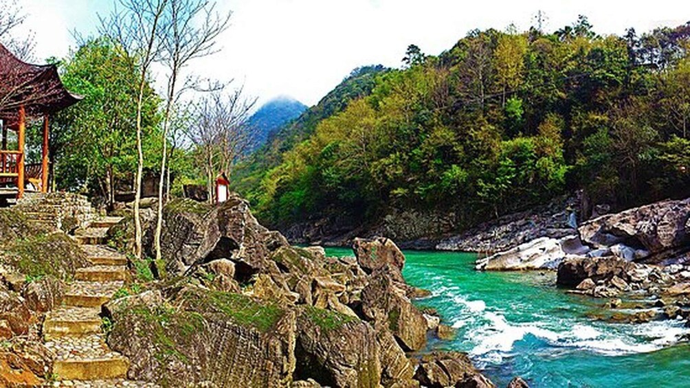 A river with rocky banks, a wooden structure with a red roof, and a forested hillside. 1