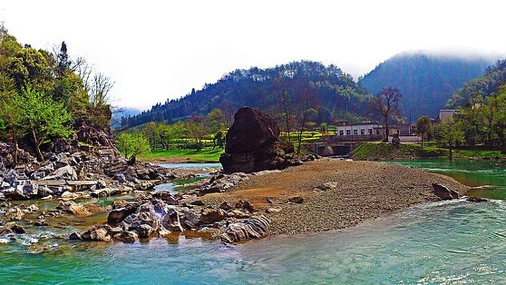 A river with rocks, a large boulder, and a building in the background. 2