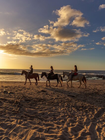 Three people riding horses on a beach at sunset. 2