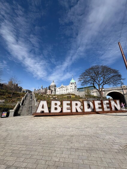 A large sign reading 'ABERDEEN' in a paved area with a staircase leading up to a hill, a tree, and historic buildings in the background. 2