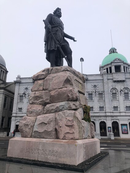 A statue of a historical figure on a pedestal with inscriptions, set against a backdrop of a large building with a green dome. 1