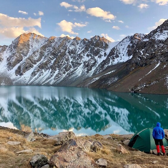 A person standing near a tent by a mountain lake with snow-capped peaks in the background. 2