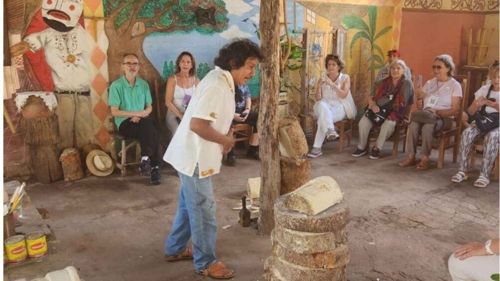 A man speaking to an audience in a rustic setting with a mural and wooden structures. 4