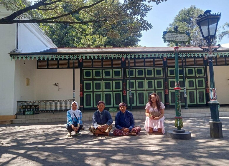 A group of people sitting on the ground in front of a building with green doors and a sign that reads 'Dilarang Foto.' 5