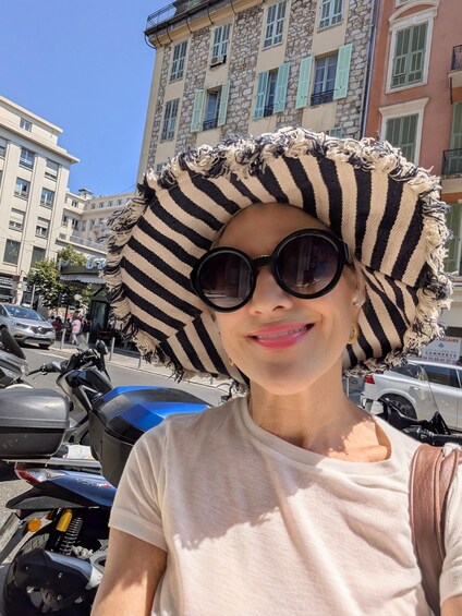 A person wearing a striped hat and sunglasses, standing in front of a building with stone facades and green shutters. 1